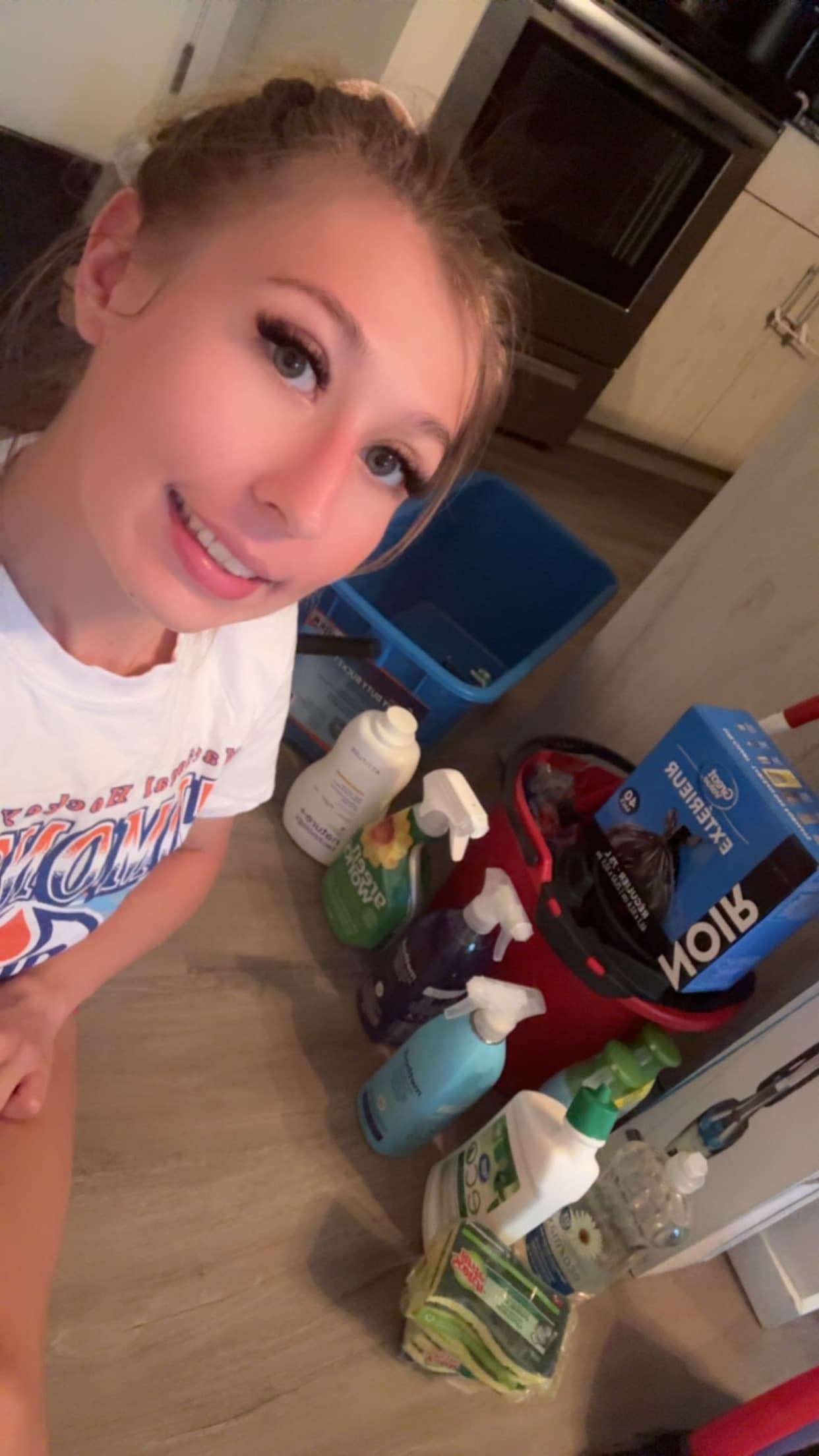 Smiling woman takes a high-angle selfie on a kitchen floor surrounded by cleaning supplies.