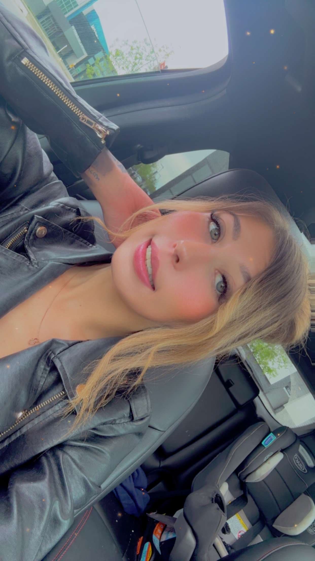 Smiling blonde woman in a black leather jacket takes a selfie inside a car.